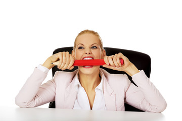 Angry business woman sitting behind the desk and biting huge pen