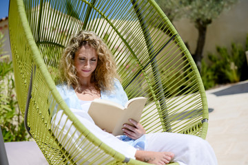 cheerful young woman reading in a hammock by the pool on sunny summer day