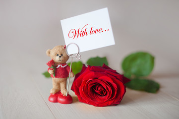 Toy teddy bear with a note with love and red rose on a light background