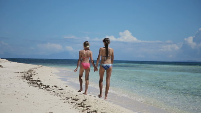 Happy sisters having fun walking on the tropical beach.Two young girls in swimsuit walking on tropical beach holding hands.Travel concept.Family,summer vacation.