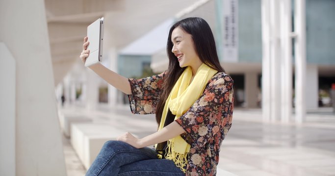 Happy single adult female in yellow scarf and flowery blouse sitting outside on bench taking a self portrait with tablet computer