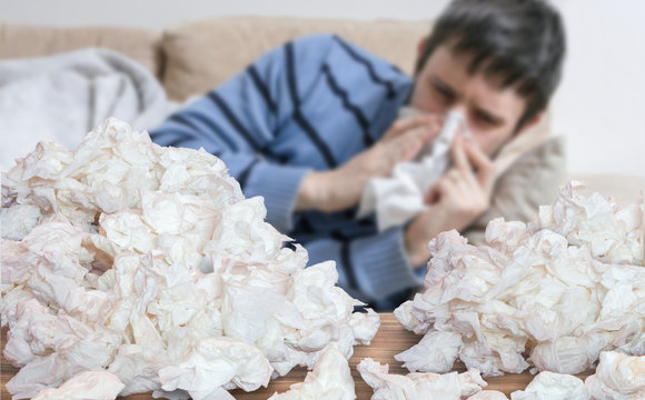 Funny sick man who has flu or cold is blowing his nose. Pile of tissues on table.