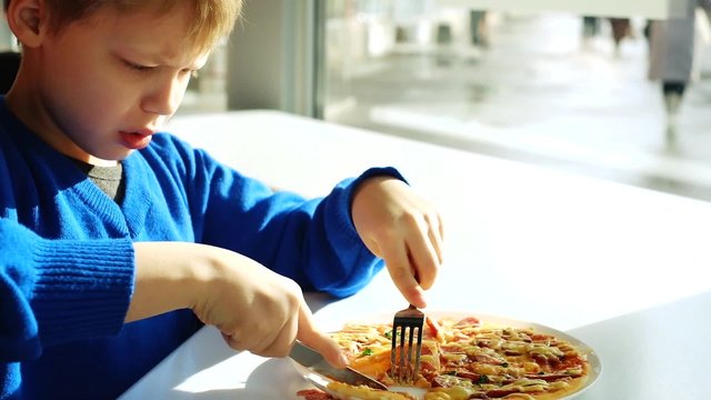 Little kid dressed in blue sweater sitting at table in fast food restaurant near window and eating pizza with fork and knife. Funny boy learning to cut food with knife. Video of eight years old boy.