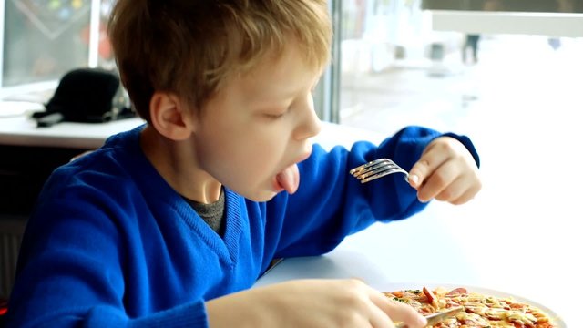 Little kid dressed in blue sweater sitting at table in fast food restaurant near window and eating pizza with fork and knife. Funny boy learning to cut food with knife. Video of eight years old boy.