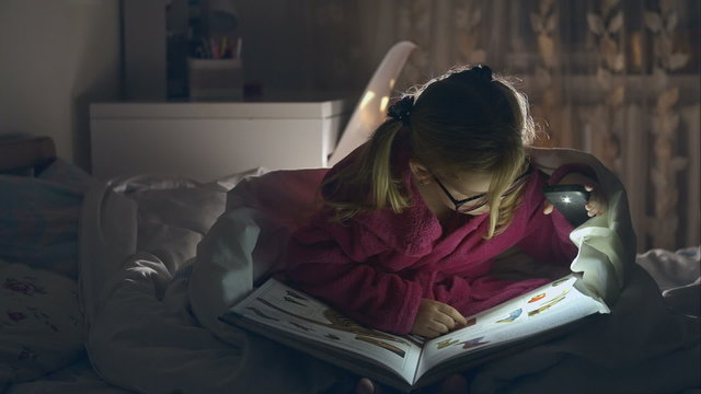 The little girl with glasses reading a book sitting in bed under the covers. Big pleasure. RAW video record.