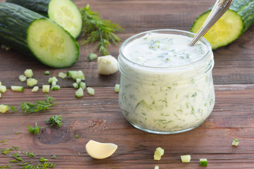 Tzatziki – traditional greek sauce in glass jar and ingredients – cucumber, garlic, dill on wooden dark background