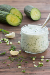 Tzatziki – traditional greek sauce in glass jar and ingredients – cucumber, garlic, dill on wooden dark background