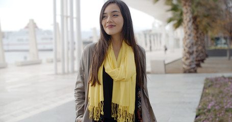 Stylish young woman enjoying a walk in town standing on a waterfront promenade looking to the side over her shoulder with a smile  wide angle close up with copy space.