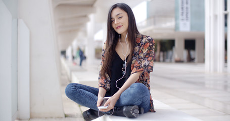 Trendy young woman listening to music on her earphones in town sitting cross legged on a bench in a pedestrian walkway with a smile of pleasure.