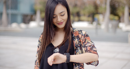 Young woman looking at the time on her wristwatch with a smile as she stands outdoors in an urban...
