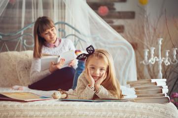 Portrait of a smiling young cute mother and daughter reading a book lying and relax on the floor in a bright big white room next to the window