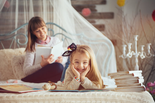 Portrait of a smiling young cute mother and daughter reading a book lying and relax on the floor in a bright big white room next to the window