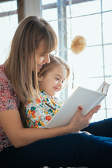 Portrait of a smiling young cute mother and daughter reading a book lying and relax on the bed in a bright big white room 