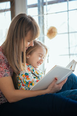 Portrait of a smiling young cute mother and daughter reading a book lying and relax on the bed in a bright big white room 