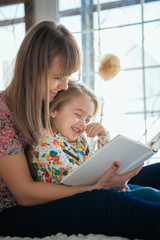 Portrait of a smiling young cute mother and daughter reading a book lying and relax on the bed in a bright big white room 