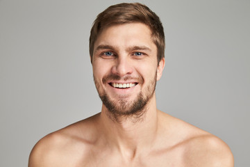Obraz premium Portrait of a smiling young man with bare swimmers shoulders on a gray background, powerful, beard, charismatic, adult, brutal, athletic, edited photo, bright smile, white teeth smile, look in camera