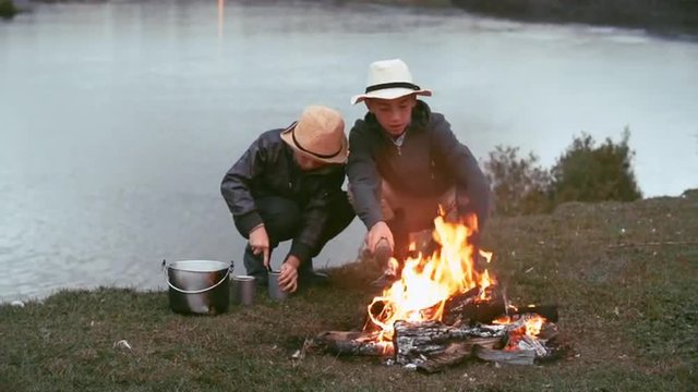 Boy making a fire on the riverbank whiles his friend opening can with food like Tom Sawyer and Huckleberry Finn during the escape from home