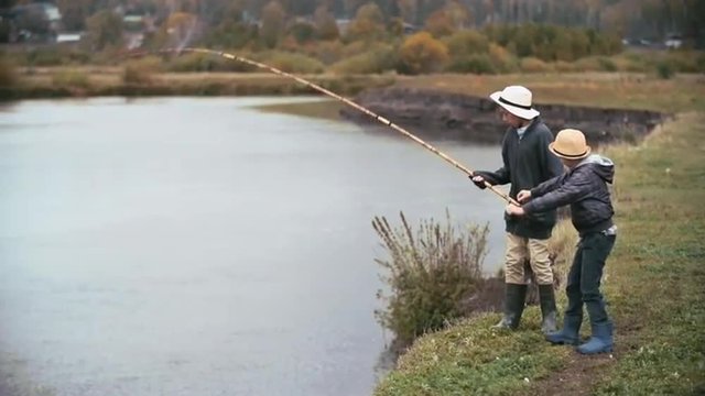 Little boy catching a big fish with rod from the river and his friend helping him to take it off from hook