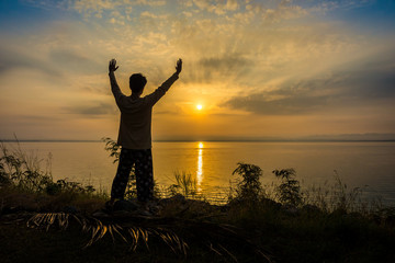 Silhouette of Asian man is refreshing by raising arms on riverside to the sunrise or sunset. Scenery of Pa Sak Chonlasit reservoir, Lopburi province, Thailand.