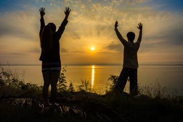 Silhouette of Asian woman and man are refreshing by raising arms on riverside to the sunrise or sunset. Scenery of Pa Sak Chonlasit reservoir, Lopburi province, Thailand.