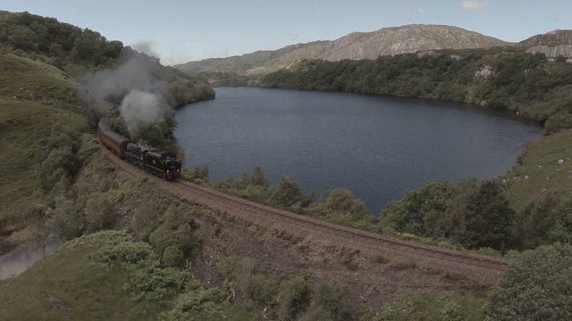Aerial shot of the Jacobite Steam Train passing a loch in the Scottish highlands en route to Fort William
