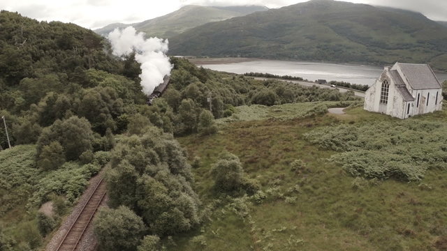 Stunning aerial shot of the Jacobite Steam Train passing a small church set among stunning scenary in the Scottish highlands
