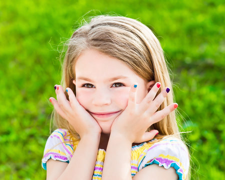 Adorable smiling blond little girl with long hair and many-colored manicure, outdoor portrait on green grass background