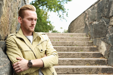 An attractive man standing, leaning against a stone wall wearing a yellow jacket outside on a sunny...