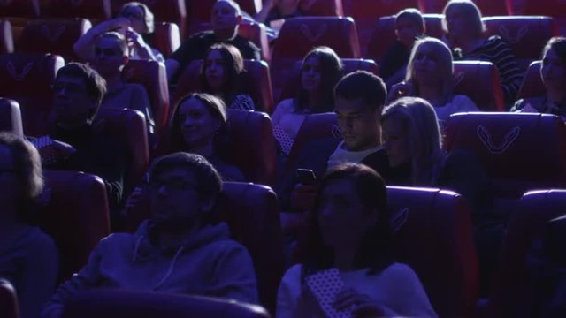 Man is using his smartphone while people are watching a film screening in a movie cinema theater. Shot on RED Cinema Camera.