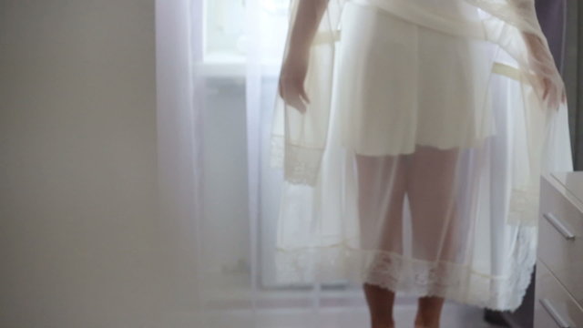 Beatful blondie bride with long sexy legs slowly takes off her white  translucent robe near the window in pale light