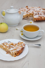 Apple pie and camomile tea set on the white wooden table decorated with fresh flowers and apples.