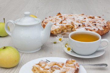 Apple pie and camomile tea set on the white wooden table decorated with fresh flowers and apples.