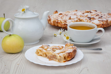 Apple pie and camomile tea set on the white wooden table decorated with fresh flowers and apples.