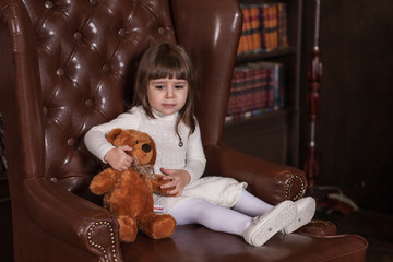 An adorable 2-year-old girl is sad. Little Princess and her toy bear sitting in a chair . Portrait in full growth in the interior