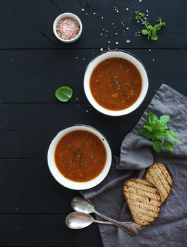 Roasted tomato soup with fresh basil, spices and bread in rustic metal bowls over black background