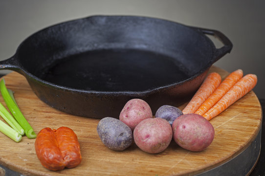 Cast iron skillet on round cutting board with small potatoes, carrots, celery & chorizo on one side with dark background