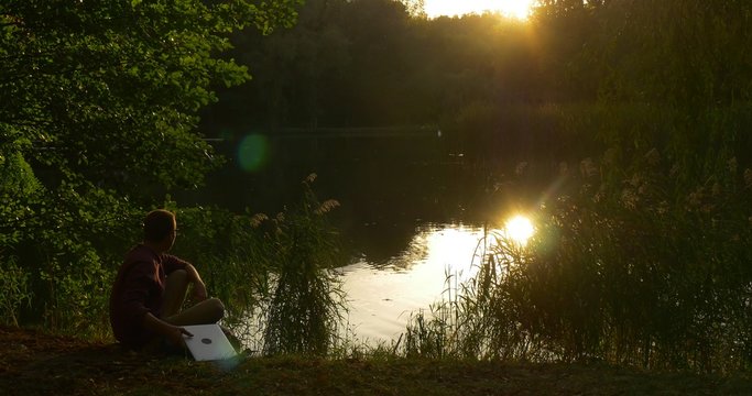 Man Sits On The Ground And Looks At The Lake He Takes Laptop And Begins To Work Programmer Copywriter Accountant Designer Freelancer Sunset Lakeshore