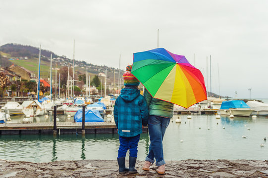 Two cute kids resting by the lake on a rainy day, hiding under big colorful umbrella, back view