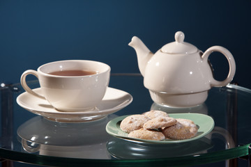 Butter lemon cookies on a green retro plate near a white ceramic cup of tea with saucer and teapot on a glass table with a blue background