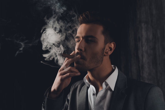 Handome confident man in suit on the grey background smoking a c