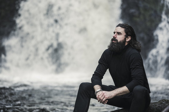 A man sitting with his hands clasped looking up, by a cascade of water, a mountain stream.