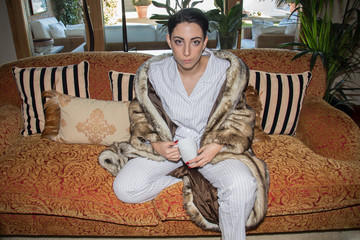 Caucasian girl with short hair sitting on the couch dressed in pajamas and fur coat. She is drinking coffee. 