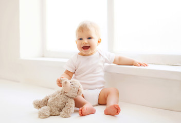 Baby with teddy bear toy sitting at home in white room near wind