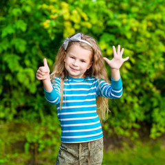 Cute blond little girl with long curly hair showing six fingers (her age) and smiling, outdoor portrait on green leaves background