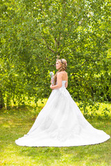 Fototapeta premium Beauty bride in bridal gown with bouquet and lace veil on the nature. Beautiful model girl in a white wedding dress. Female portrait in the park. Woman with hairstyle. Cute lady outdoors