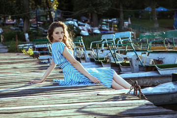 Beautiful young fashion model with freckles on her face and light blue striped dress and fashion makeup and hairstyle is sitting on pier, posing and looking into the distance.