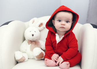 Portrait of cute adorable Caucasian smiling sad baby boy girl with black brown eyes in red hoodie shirt sitting in chair with toy looking directly in camera
