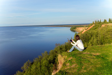 Young woman sitting on the shore of the lake with tablet compute