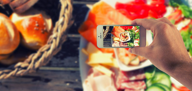 M&auml;nnliche Hand haltet ein wei&szlig;es Smartphone und fotografiert damit einen Esstisch mit Kr&auml;utern und Vesperplatte