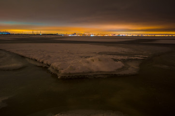 Big night city Saint-Petersburg on the horizon behind Baltic sea in spring time. Baltic sea frozen. City reflect lights in the sky.